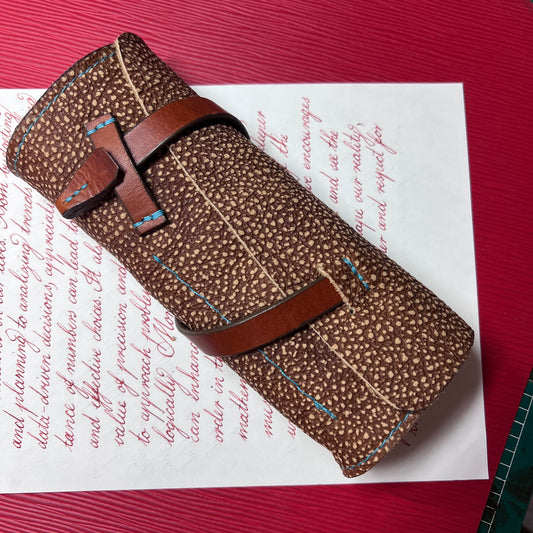 A golden brown patterned bison leather fountain pen case with a brown leather straps and blue contrast stitching, resting on a sheet of paper over a red textured surface.