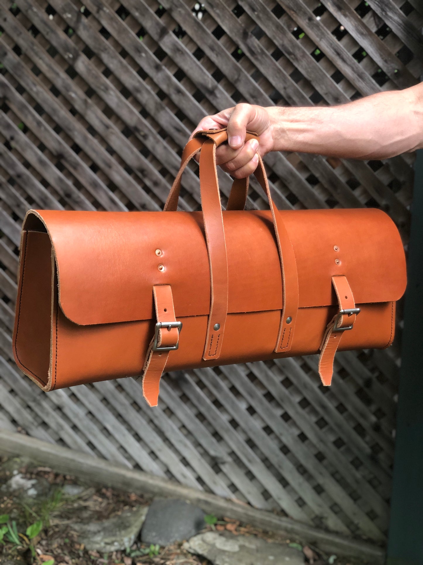 Brown leather tool bag with buckles, held by a person against a wooden lattice background