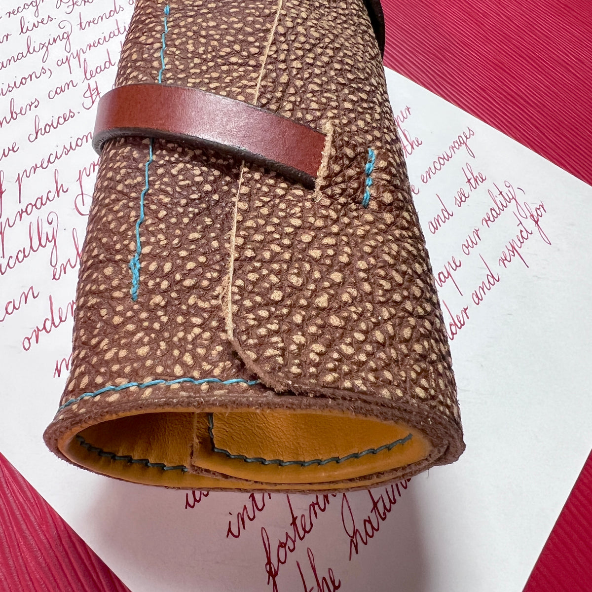 A close-up of a rolled brown textured leather pen roll, pen wrap sleeve or pouch with a darker brown strap and visible blue contrast stitching, placed on white paper with red script over a red surface.