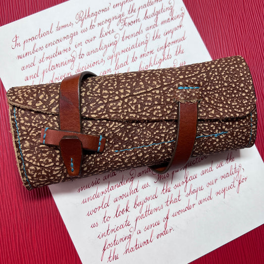 A brown patterned leather fountain pen roll with a brown leather strap and buckle closure, laid diagonally on white paper with red cursive writing over a red textured background.