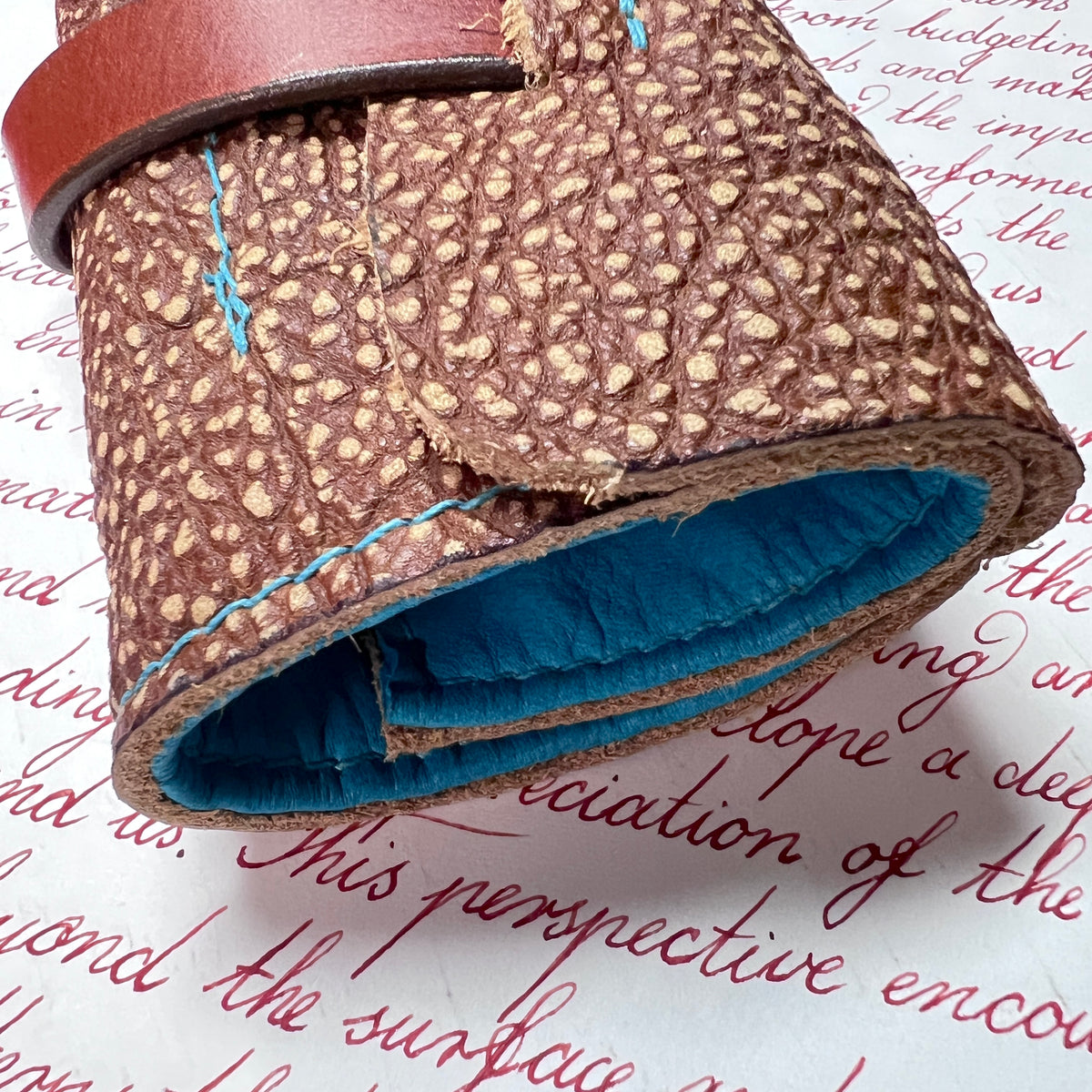 A close-up of a rolled brown textured leather pen case, pen roll, a pouch or sleeve, showing a blue deerskin lining and light blue stitching, placed on a white paper background with red cursive writing.