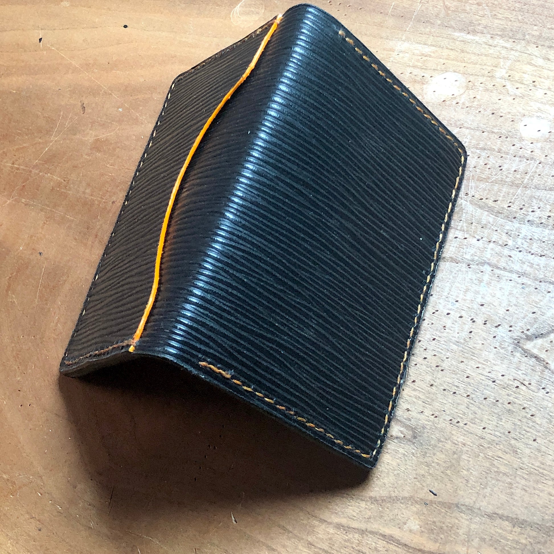 Black textured wallet with yellow stitching on a wooden surface. EPI leather luxury wallet, card case with goatskin leather lining, hand stitched, finished edges. 