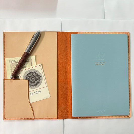 An open leather planner or notebook cover in warm tan and orange-brown tones, shown on a white background. The right side holds a light blue notebook, and the left side has a pocket holding a pen with a dark wood-look grip and two small cards.
