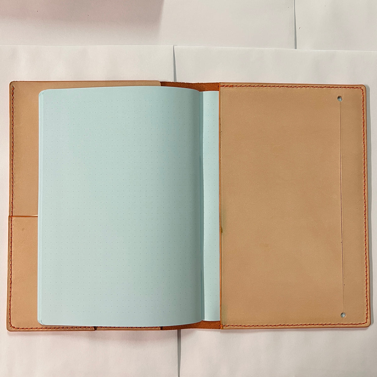 An open leather notebook cover or passport holder with tan interior pages and a light blue sheet inserted, photographed from above on a white surface.
