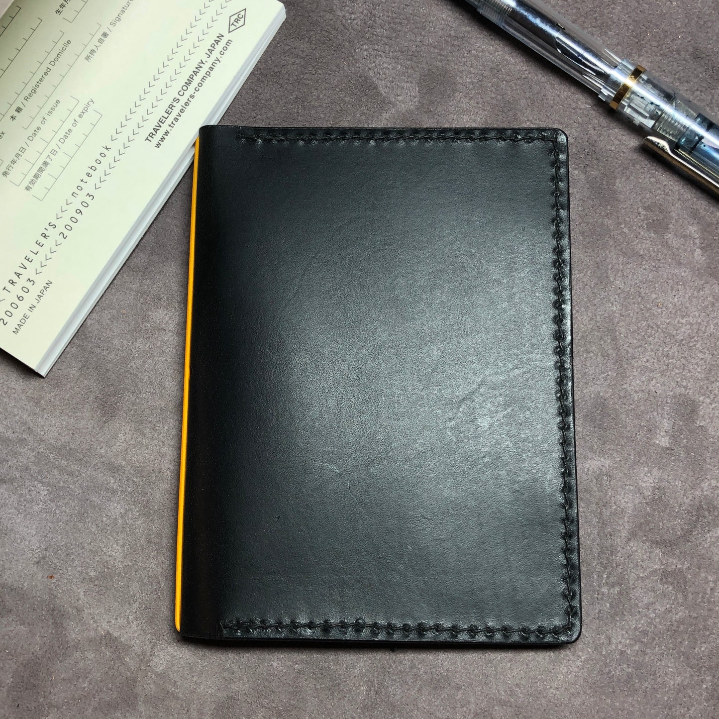 Cowhide leather Passport size Travelers Journal cover.  Hand stitched, finished edges. One Midori Travelers Notebook Journal included.  Black leather notebook with a fountain pen on a gray surface.