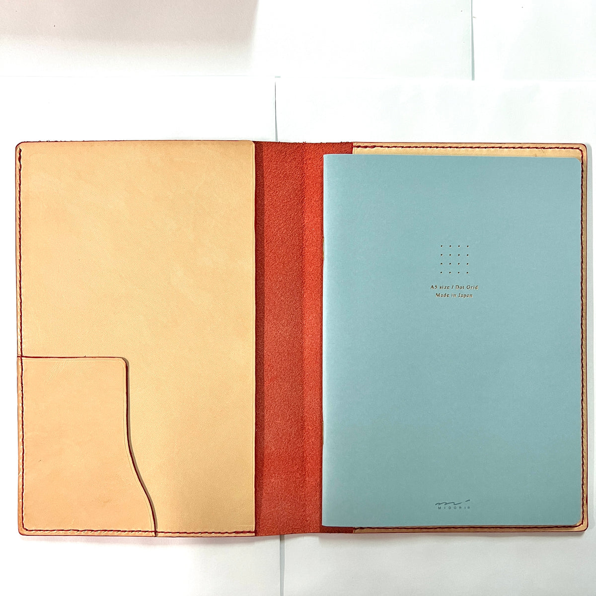 An open leather journal cover in natural tan and rust red, holding a light blue printed page with small text and a dotted grid.