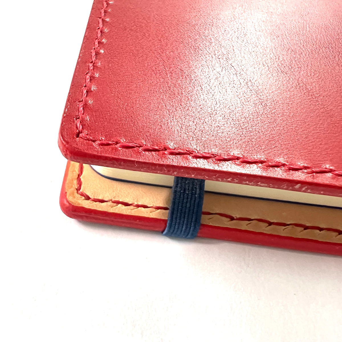 Close-up of a red leather notebook cover with visible stitching, a tan interior page edge, and a dark blue elastic closure band on a white background.