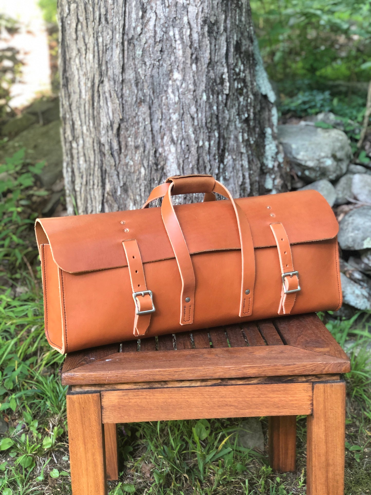 Brown leather tool bag with buckle closure on a wooden bench with a natural background.