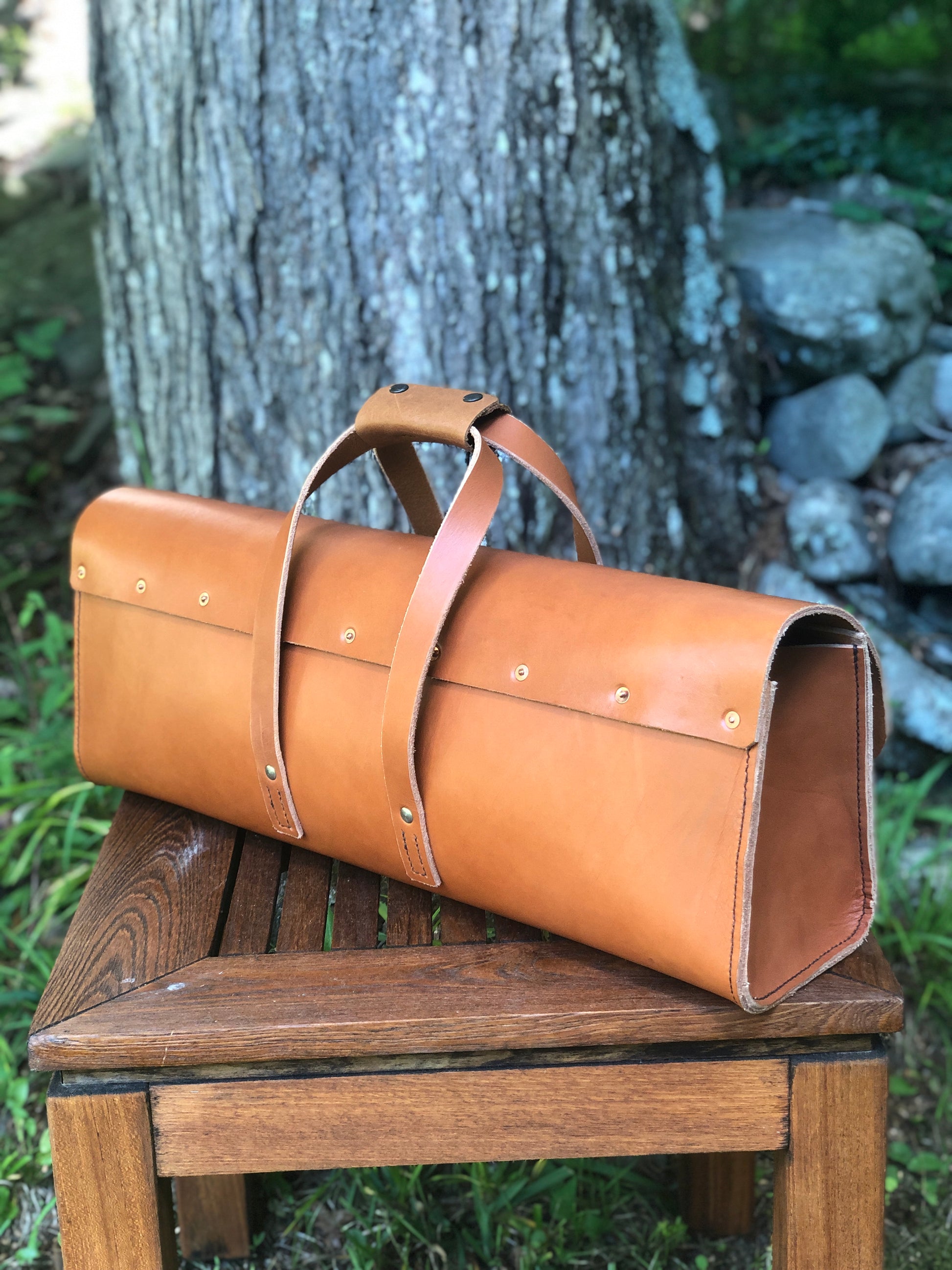 Brown leather tool bag with buckle closure on a wooden bench with a natural background.