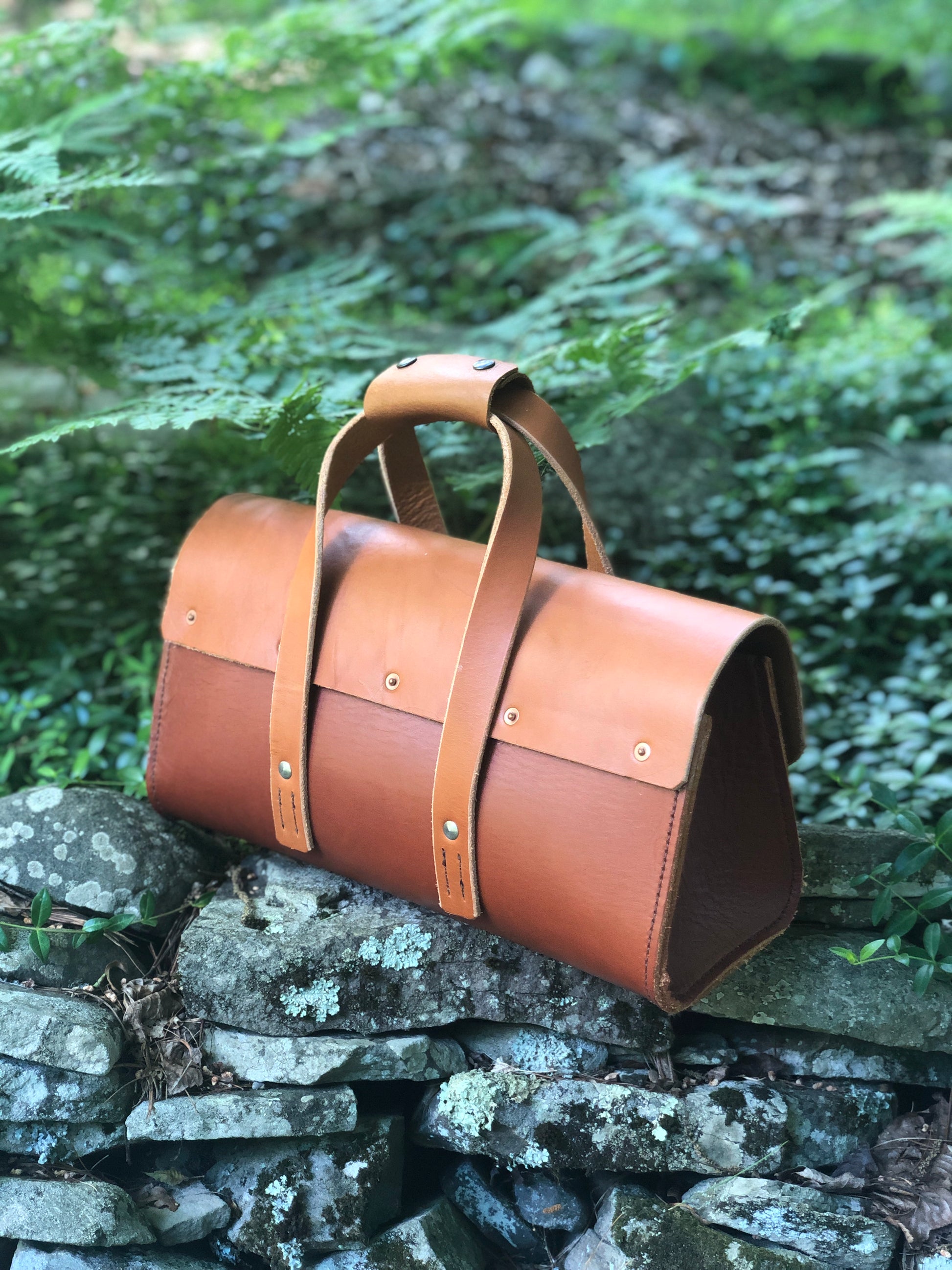 Whiskey and Chestnut brown leather tool bag, roll-up bag on a stone wall with greenery in the background