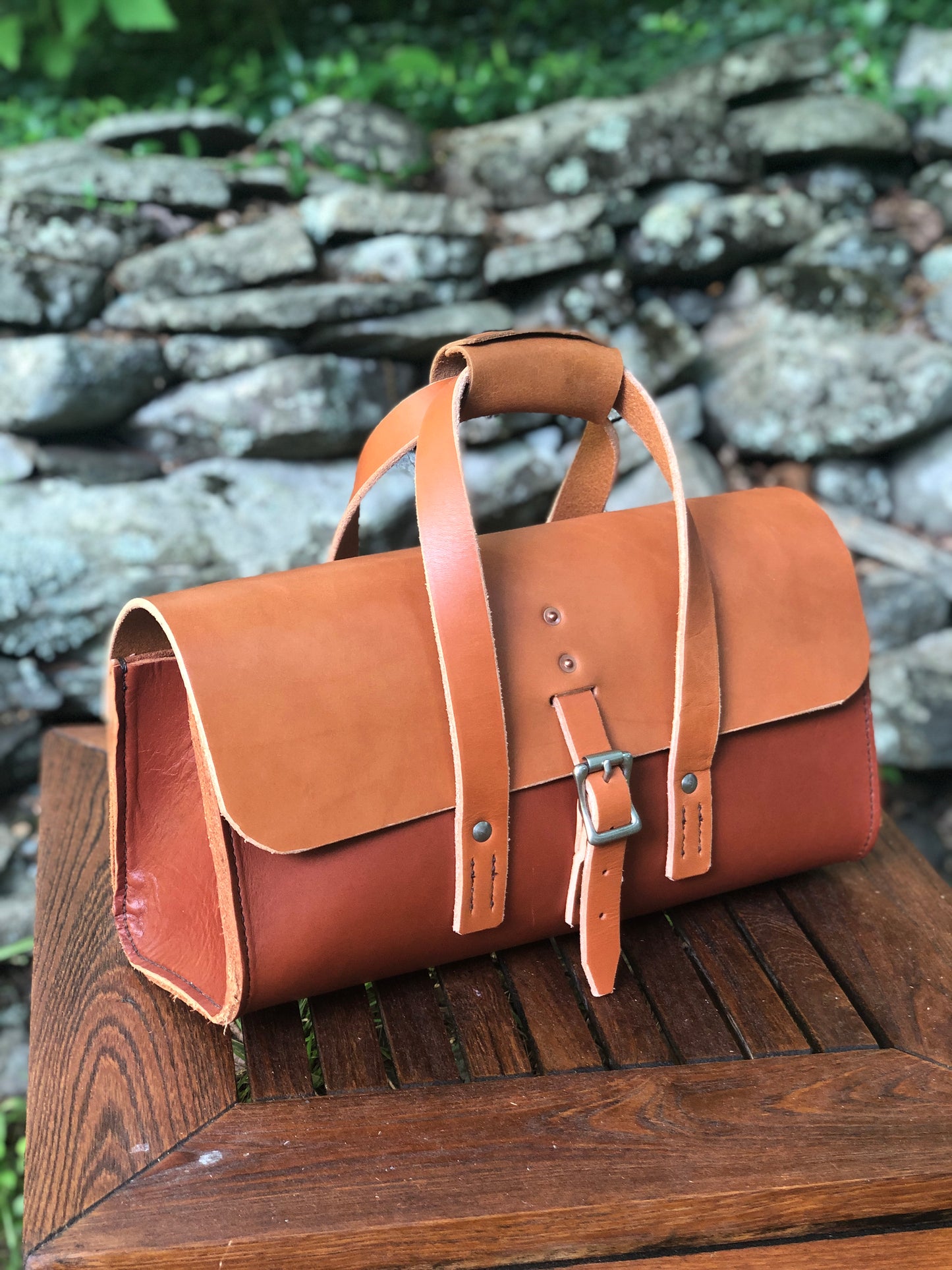 Whiskey and chestnut brown leather tool bag on a wooden surface with a stone wall background