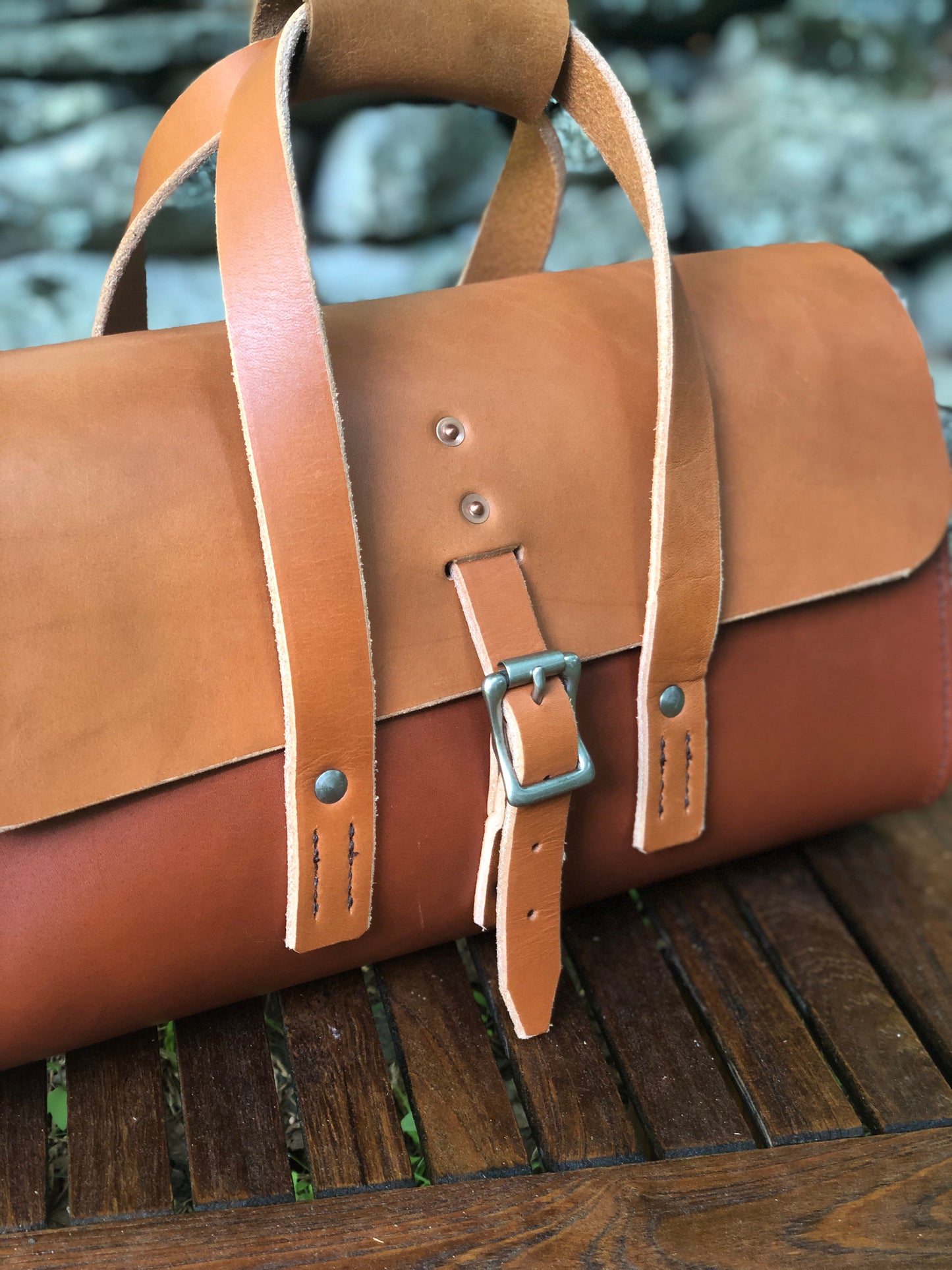 Whiskey and chestnut brown leather tool bag on a wooden surface with a stone wall background.
Brown leather tool bag with closure on handles, strap on a wooden surface