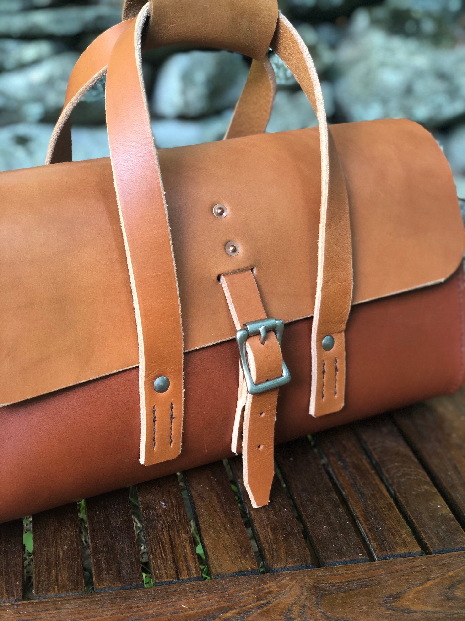 Whiskey and chestnut brown leather tool bag on a wooden surface with a stone wall background.
Brown leather tool bag with closure on handles, strap on a wooden surface