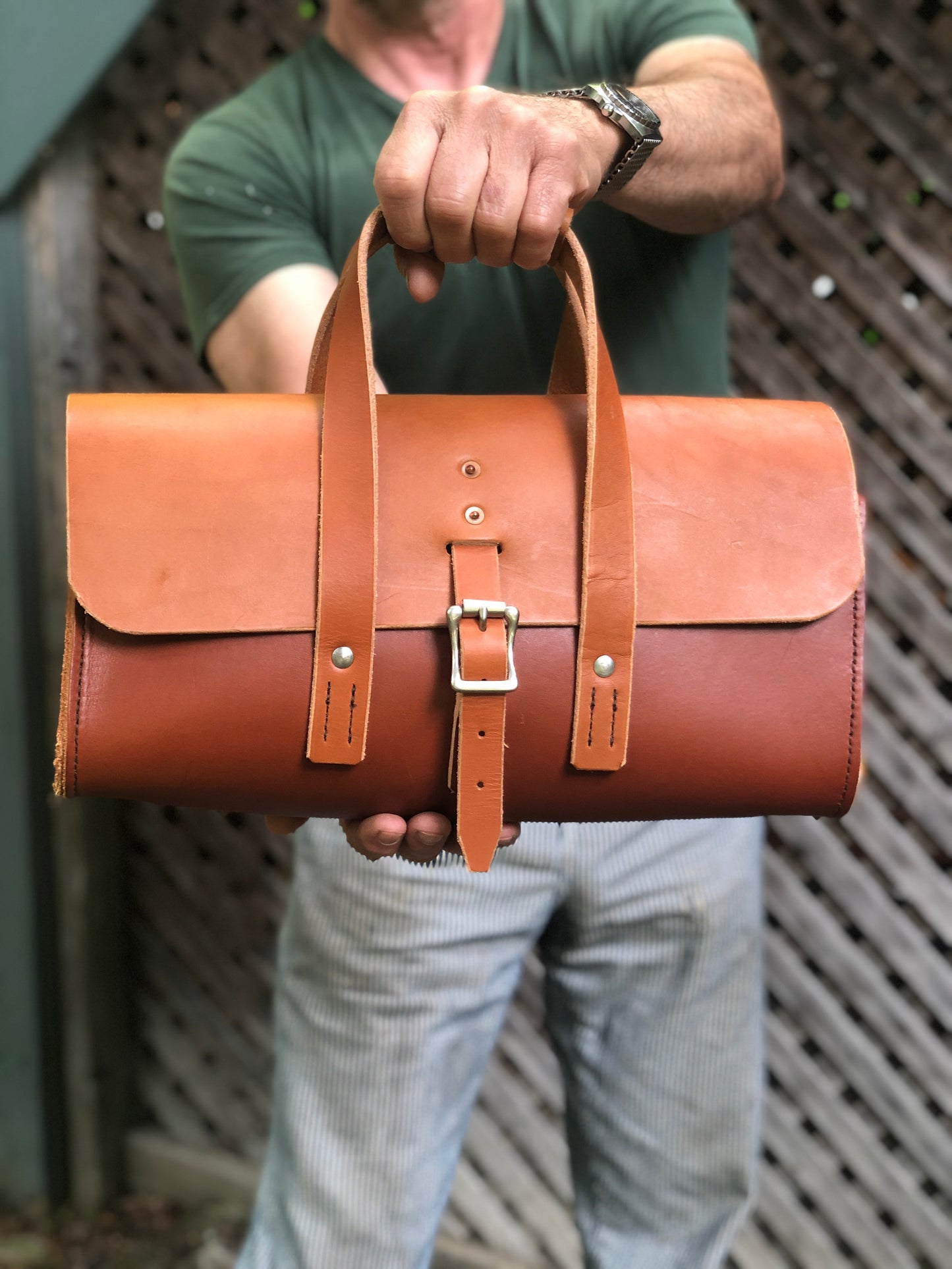 Whiskey and chestnut brown leather tool bag on a wooden surface with a stone wall background.
Person holding a brown leather tool bag against a wooden background.