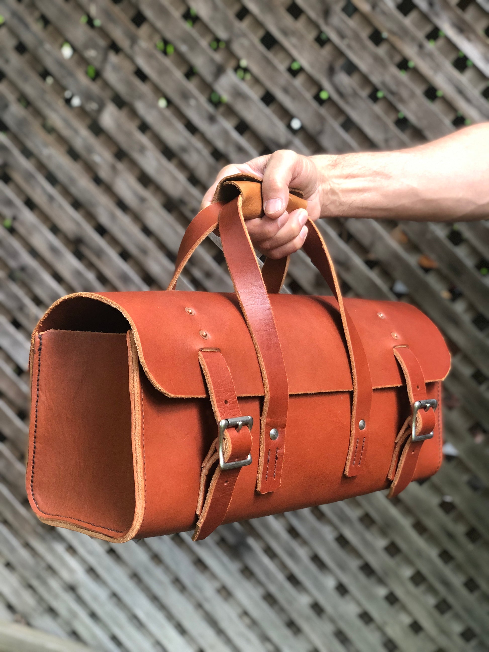 Brown leather tool bag, satchel bag held by a hand against a metal grid background